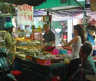 Cơm Tấm stall on the street. A board titled "Cơm Tấm" is hung on top of the stall. On the stall, a cook is making Cơm Tấm for his customers who are waiting around the stall for their to-go order. In front of him is a small, transparent glass cupboard where he keeps his ingredients ready to put in the dish. Behind the stall, it looks like a small building where this stall stores its stuff. Most of the time, in Vietnam, this "building" is the owner's house. This is how people do casual Cơm Tấm business in Vietnam.