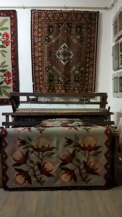 A carpet with a geometric ornament attached to a wall, and a loom with a nearly finished carpet with a floral ornament