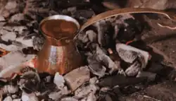 A small metal pot filled with coffee resting on hot coals