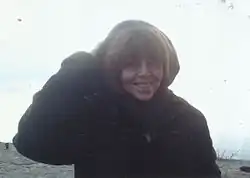 blurry image of Shlomit C. Schuster wearing a dark coat with a hood, grinning directly at camera, with body of water in background