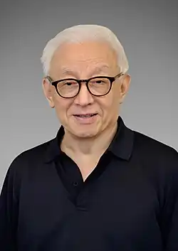 Man with white hair and dark rimmed glasses wears a white dress shirt, dark dress pants, and a navy blue bullet resistant vest while sitting in a beige chair with light wood trim