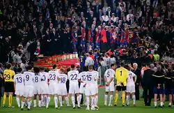 A line of men in white shirts watching men in blue-and-red shirts standing on a rostrum in the background.
