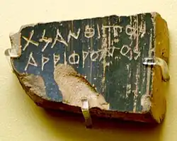Color photograph of a piece of pottery, damaged in three places, with two lines of Greek words