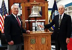An interior wooden display case with a brass ship's bell flanked by two men in coat and tie standing on either side.