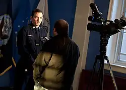 A reporter interviews another man. A camera is set up to the man's right in the picture. The reporter holds a microphone with a "Fox CT" mic flag.