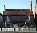 Front view of two joined flint two-storey cottages with white wooden doors next to each other under a shared tiled hood. There are two ground-floor bow windows and two tall rectangular windows above. The red-tiled roof has chimneys at each end; the right-hand one is much taller, while the left-hand one is painted white. There is a low white fence in front.