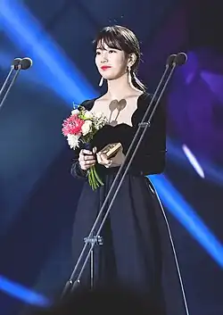 Bae Suzy wearing a long black dress on stage, smiling while holding a bouquet and an award