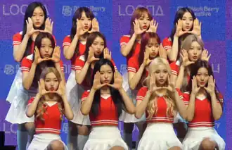 Twelve young women all wearing identical red short sleeved knitted crop tops and white tennis skirts standing in a 4 by 3 configuration with their hands up to their faces in a 'shouting' gesture. The four girls in the top row are standing straight, while the four girls in the middle row are leaning forward slightly, and the four girls in the bottom row are kneeling on one knee.