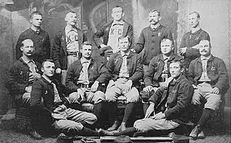 Baseball players are posing for a photograph, five men standing, five men sitting on chairs, and two are sitting on the floor.
