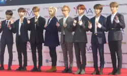 Eight men stand in a row, facing right and waving or holding peace signs