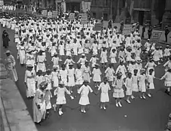 A large group of African American children dressed in white clothing, marching in a parade on a wide street