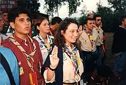 Outdoors, a group of about 10 young people wearing scouting scarves and other insignia with several showing the three finger scout salute