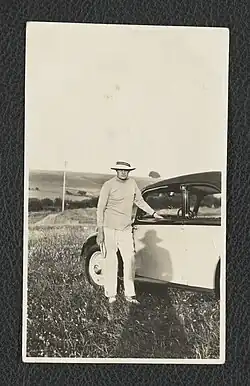 A black and white portrait photograph of a man in a hat, a light coloured long sleeve jumper, and light coloured trousers. He is leaning against a car door, and holding a notebook in his hand.