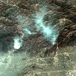 An aerial image from high above the fire shows gray smoke emanating from a brown burn scar in rugged mountains, with dense suburbs clustered on their southern extent