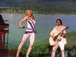 A blond female dressed in a white leotard with purple stripes, stands in front of the image of a tropical island. To her right, there's a man dressed in white playing guitar.