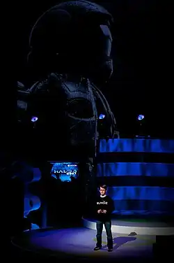 A man stands on a large stage, talking to the audience. Behind him is a flatscreen television displaying the game's title menu; behind that is a large representation of an ODST.