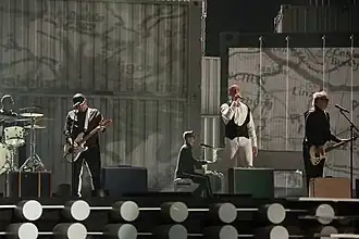 Five men performing on a stage, with four of them playing instruments.