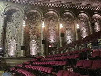 The theater's balcony and right wall. The balcony has red seats on a slope, and the wall has several arches.