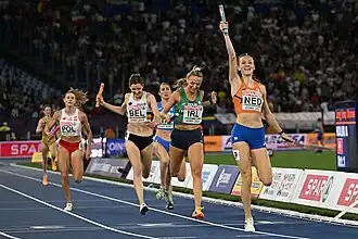 Photo of Femke Bol raising her right hand holding the baton as she finishes the relay race followed by five other runners