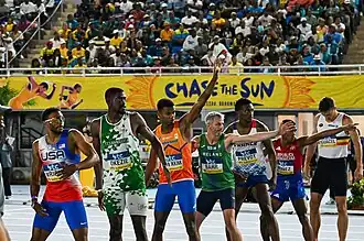 Photo with in the front seven sprinters waiting to receive the batons, some holding up their arms, and in the background spectators sitting in stadium seats