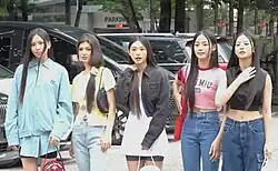 Five women standing outdoors, dressed in casual outfits, each with long, straight hair. Behind them are parked vehicles and some trees.