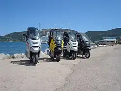 Four BMW C1 scooters parked side by side in front of a lake, with mountains and the masts of a large ship in the background