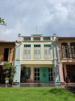 Two-storey shophouse with "five foot way" on ground floor before entrance doors, three windows with wooden shutters on second floor, and ornamental green ceramic latticework on the top of the facade. A flagpole extends from the top. There is a grassy lawn in front of the building.