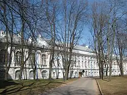 A long, white and light-blue classical building is seen from a distance through a stand of bare, tall trees. A paved road leads through the trees towards an entrance in the center-right of the building's facade. The sun is out, casting long shadows of the trees across a grassy lawn in the foreground under a clear blue sky.