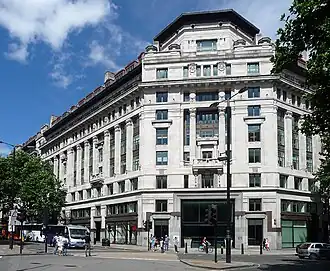A modern colour photograph of a large white building in central London, taken during sunny weather. The background is a blue sky, pedestrians can be seen at the bottom of the photograph.