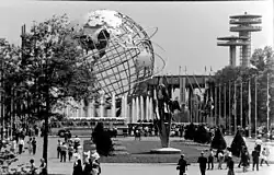 A huge, skeletal, globe-like structure representing Earth dominates a concrete plaza with trees and a bus, and a futuristic building rises in the background.
