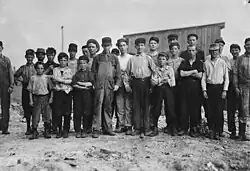 20–25 males facing camera in a row, mostly children. Ages vary. They are in work clothes. Some are dirty. Some wear caps. They stand on dirt with a wooden building behind them.