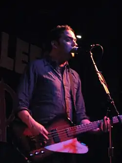 Adam Schlesinger is playing a guitar and singing live in front of a microphone.