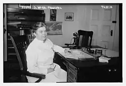 A middle-aged white woman in a nurse's uniform, seated at a desk