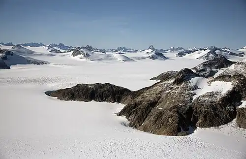 A distance shot near Juneau, Alaska. The area is covered in untouched white snow with mountain peaks in the distance. To the right of the image, nearer the photographer, is an uncovered outcrop of rocks.