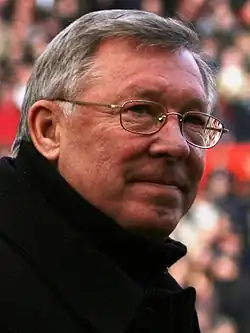 Photograph of Alex Ferguson. He is wearing a dark coat with a high collar and a pair of spectacles, and is looking off to the left