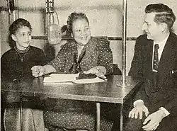 A photograph of two women and a man seated at a table with a hanging microphone in the center and papers on the table