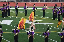 Two color guard members dressed in togas spin flags with fire designs in the middle of the DVC football field. Surrounding these color guard members are flute and saxophone players standing still and playing. They are dressed in a purple jacket and black marching pants and are wearing shakos.