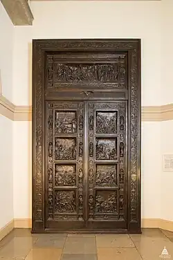 A brown bronze door between two grey walls. The door has eight indents, two on each side. The door is engraved with small dioramas.
