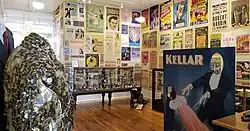 Photo of a well lit room with a glass case of magic paraphernalia, a sparkling magician's suit, and a photo stand-in where you can pretend to be a magician levitating a woman. On the wall are many colorful posters.