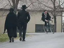 A couple in dark clothes walk along a road, backs to the camera, while a young man on a bicycle heads in the opposite direction.