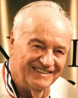 Colour press shot of an elderly, smiling man with short, receding white hair.