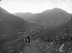 A man on a horse wearing a jacket and hat, looking out into valley from the Otto Mears road; road leads down to the town of wood-frame structures and the Gold Prince Mill. Photo circa 1888.