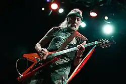 A man with long white hair playing electric guitar on a stage.