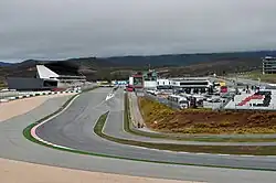 A tarmac course in hilly terrain, lined with concrete and gravel run-off areas, passes between a large grandstand and the pit garages.