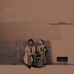 A sepiatone photograph of two Black women sitting on chairs in front of a brick wall with a "Leather Blvd" street sign above them.