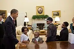 Barack Obama pointign at the call button on the Resolute desk while four children look on.