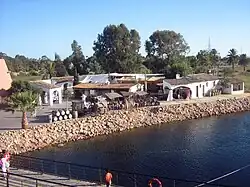 Colour photo showing the reconstruction of the ancient port of Palos de la frontera: transportation barrels, low buildings bordered by a covered gallery, a chariot at the very edge of the water awaiting the arrival of a boat.