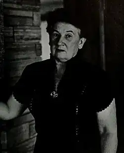 Portrait of a middle-aged woman in a short-sleeved shirt adorned with a large brooch standing next to a bricked rock wall