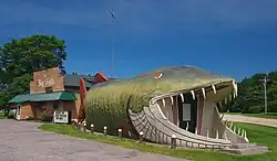 65-foot long wooden structure shaped and painted like a giant fish, flanked by a log cabin structure with sign reading "The Big Fish"