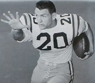 Black and white posed shot of Cannon from the thighs up, in the stiff-arm position, cradling a football, wearing pads and a jersey number 20, helmetless